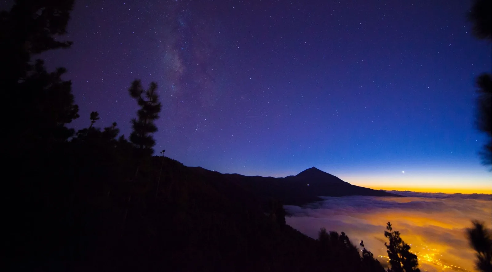 Teide's silhouette at blue hour above a sea of clouds glowing with town lights below