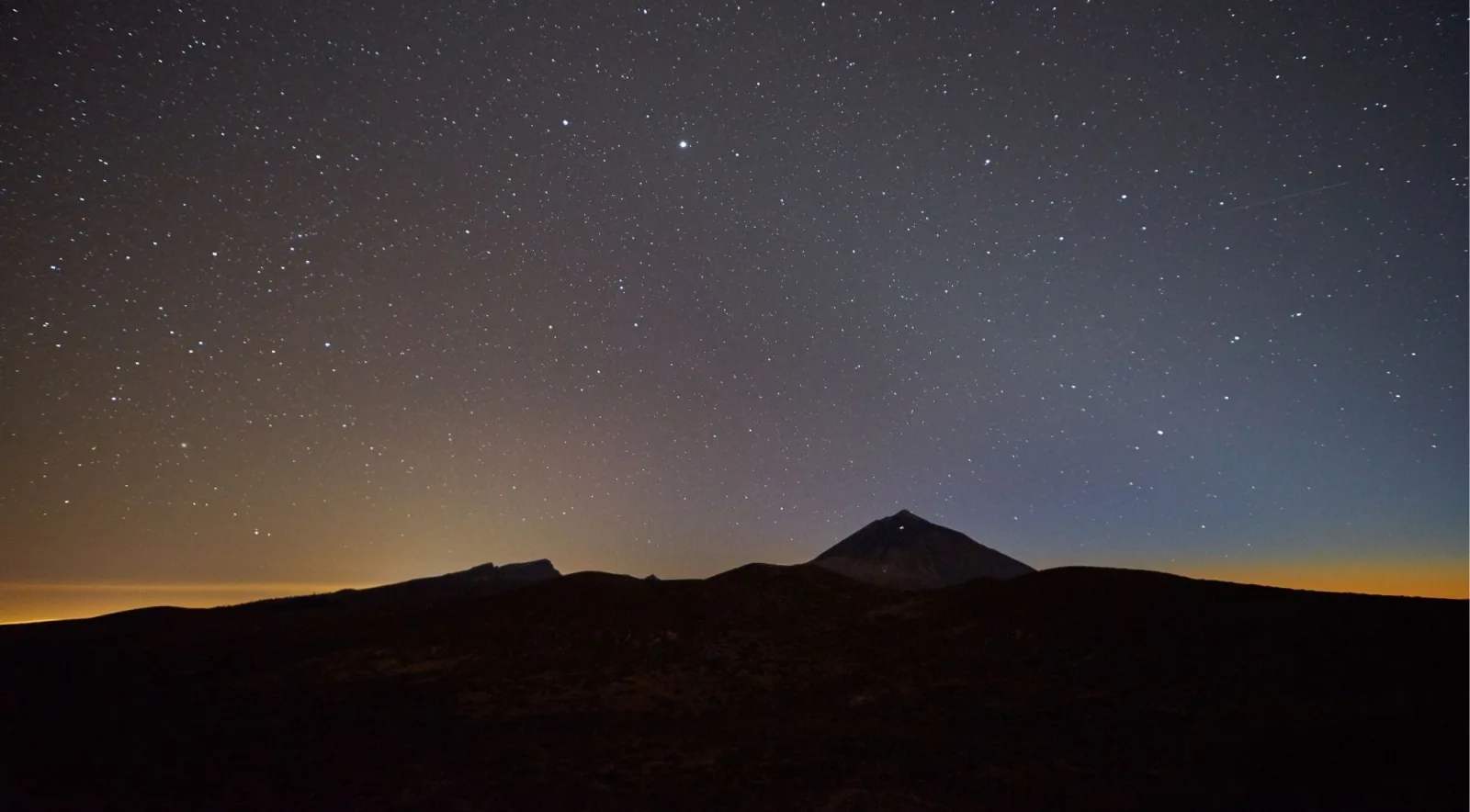 A dense field of stars above the dark outline of Mount Teide with airglow on the horizon