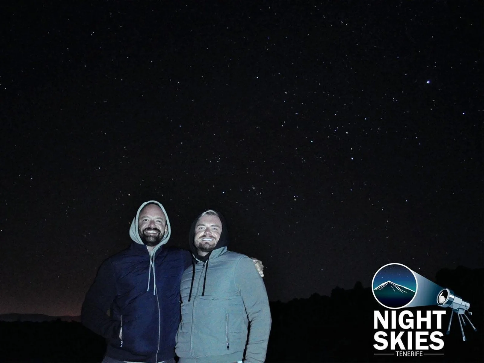 Two guests in hooded jackets standing under a black, star-filled sky